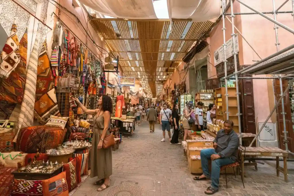 Tourists browsing colorful rugs and crafts in a narrow souk street in Marrakech, Morocco